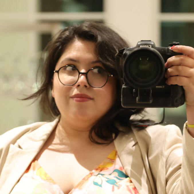 Latino woman in formal attire, named Val, is holding a full-frame DSLR camera.